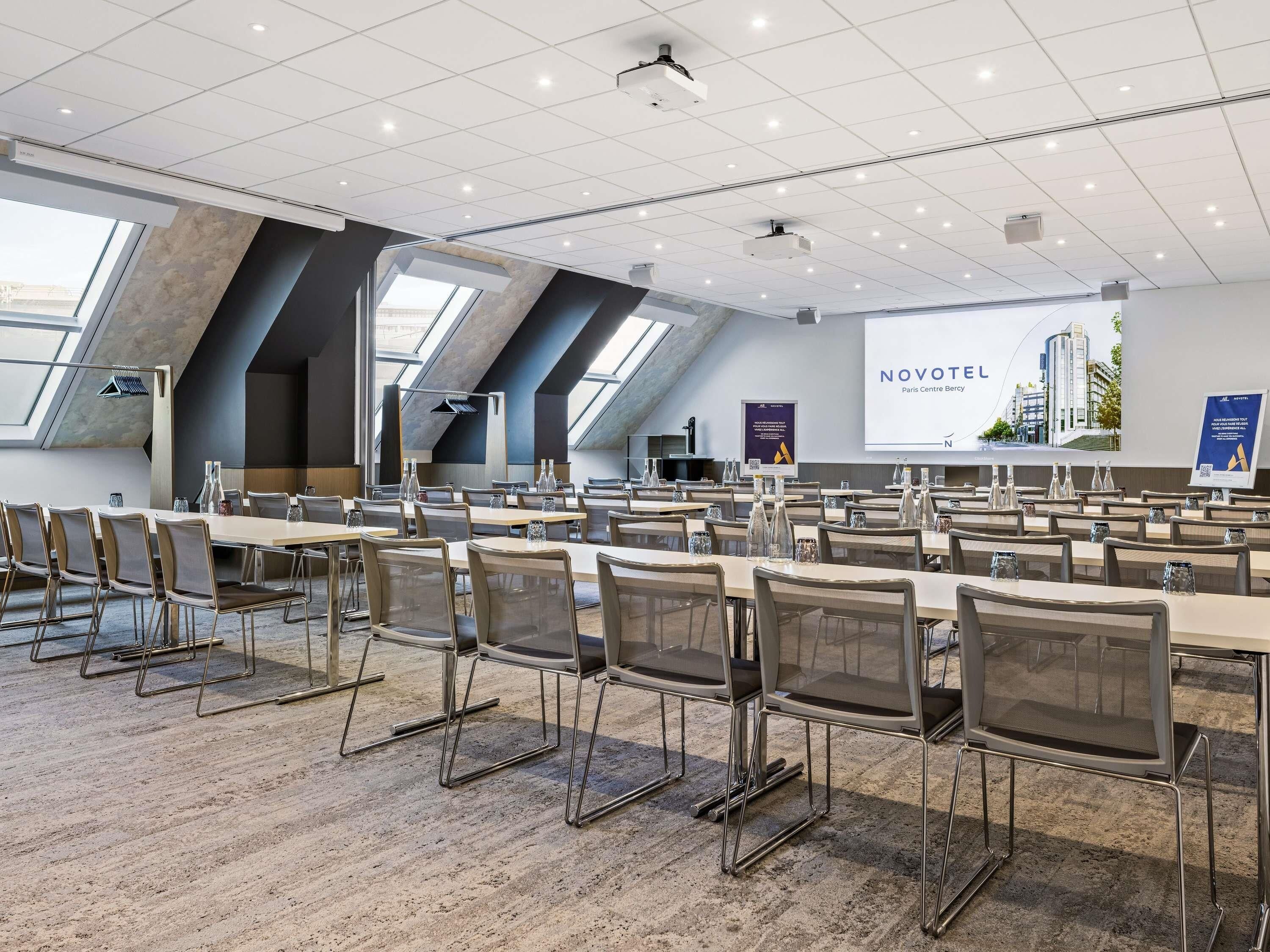 A conference room with multiple tables and chairs, a projector screen, and a large window with a view of buildings.