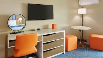 Een hotelkamer met een wit bureau, oranje stoelen, een ronde spiegel, een flatscreen televisie, een lamp en een klein tafeltje.