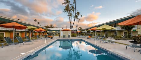 Una piscina con sillas de descanso y sombrillas, rodeada de palmeras y edificios de hotel.