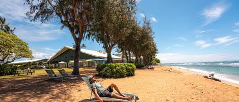 Escena de playa con una persona recostada en una silla de descanso bajo un árbol, un edificio con techo verde al fondo y el océano en la distancia.