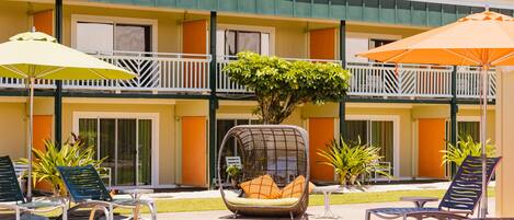 A pool area with lounge chairs and umbrellas, a small tree planter, and a building with balconies.