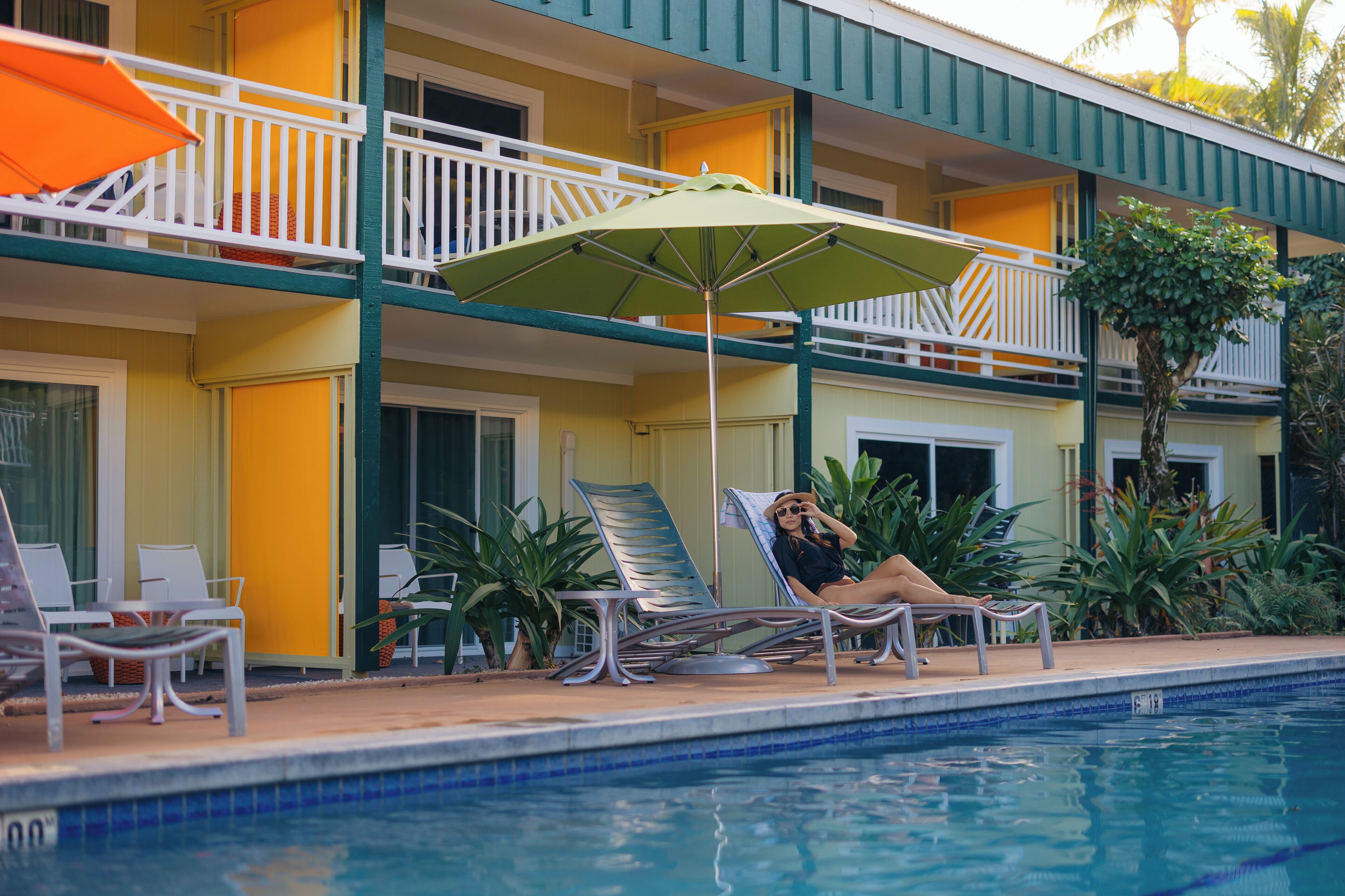 Un área junto a la piscina con sillas de descanso y una sombrilla, un edificio con balcones y una piscina.