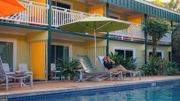 Un área junto a la piscina con sillas de descanso y una sombrilla, un edificio con balcones y una piscina.