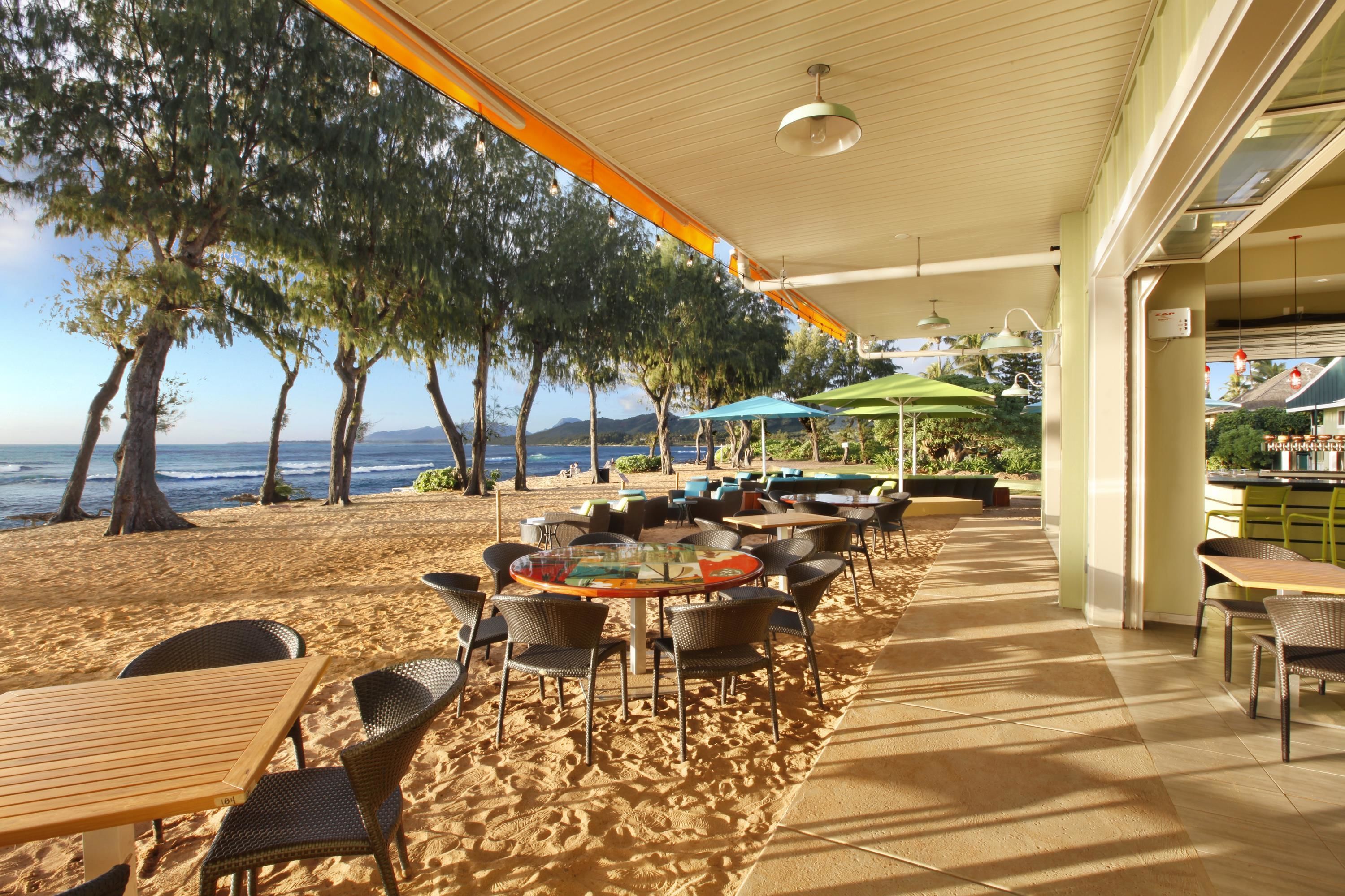 A beachside restaurant with outdoor seating, tables, and chairs, overlooking the ocean and trees.