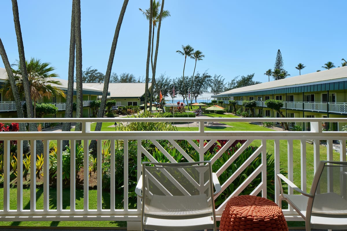 A hotel balcony with white railings, two white chairs, and a woven ottoman.