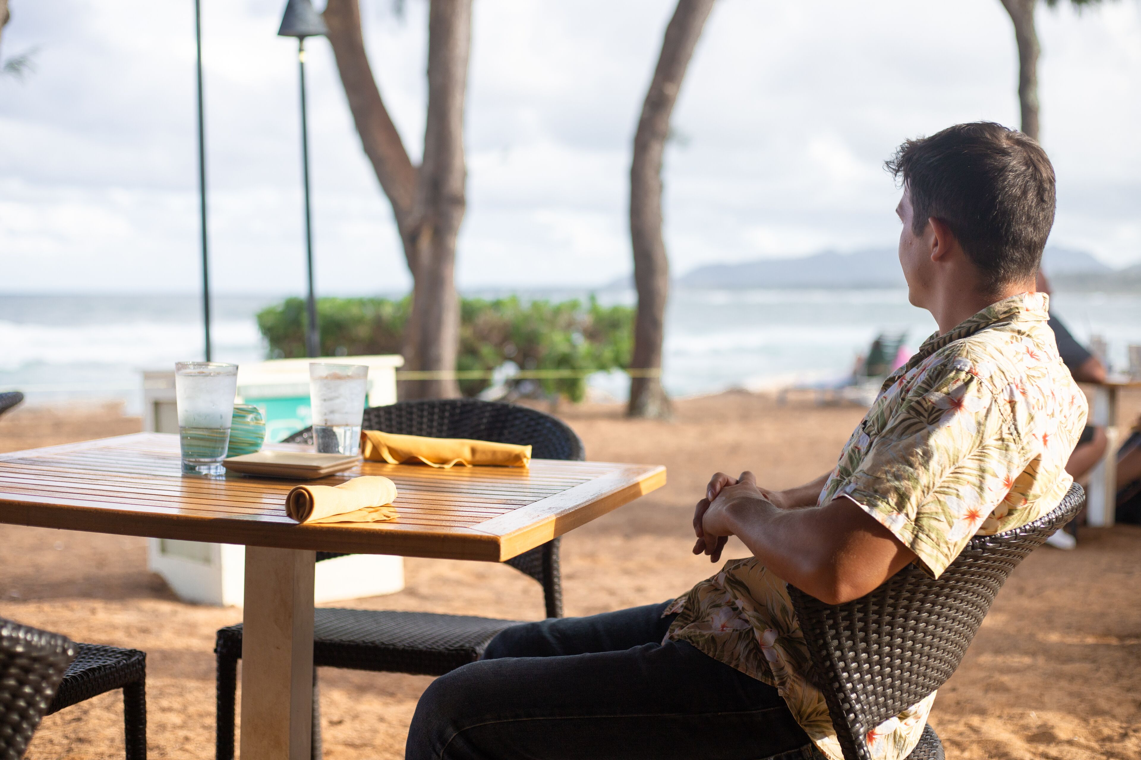 A person seated at a wooden table with a drink and a napkin, overlooking a beach with palm trees and the ocean.