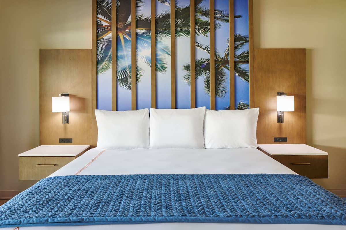 A hotel room with a large bed, two bedside tables with lamps, a wall mural of palm trees, and a blue textured bedspread.