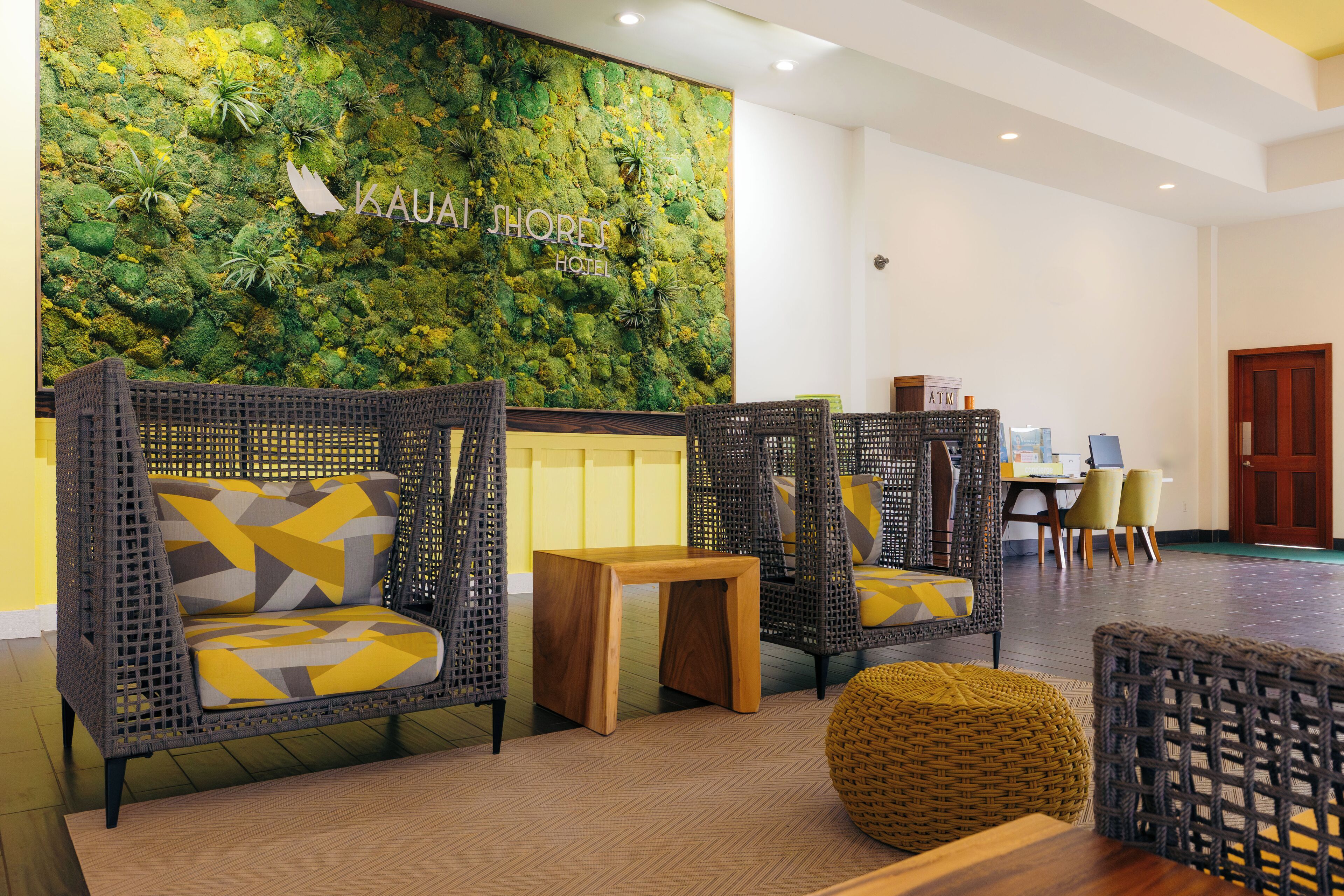 A lobby with wicker furniture, a wooden table, and a green wall with the text "Kauai Shores Hotel".
