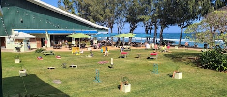 An outdoor dining area with tables and chairs, a building with a green roof, and a view of the ocean.