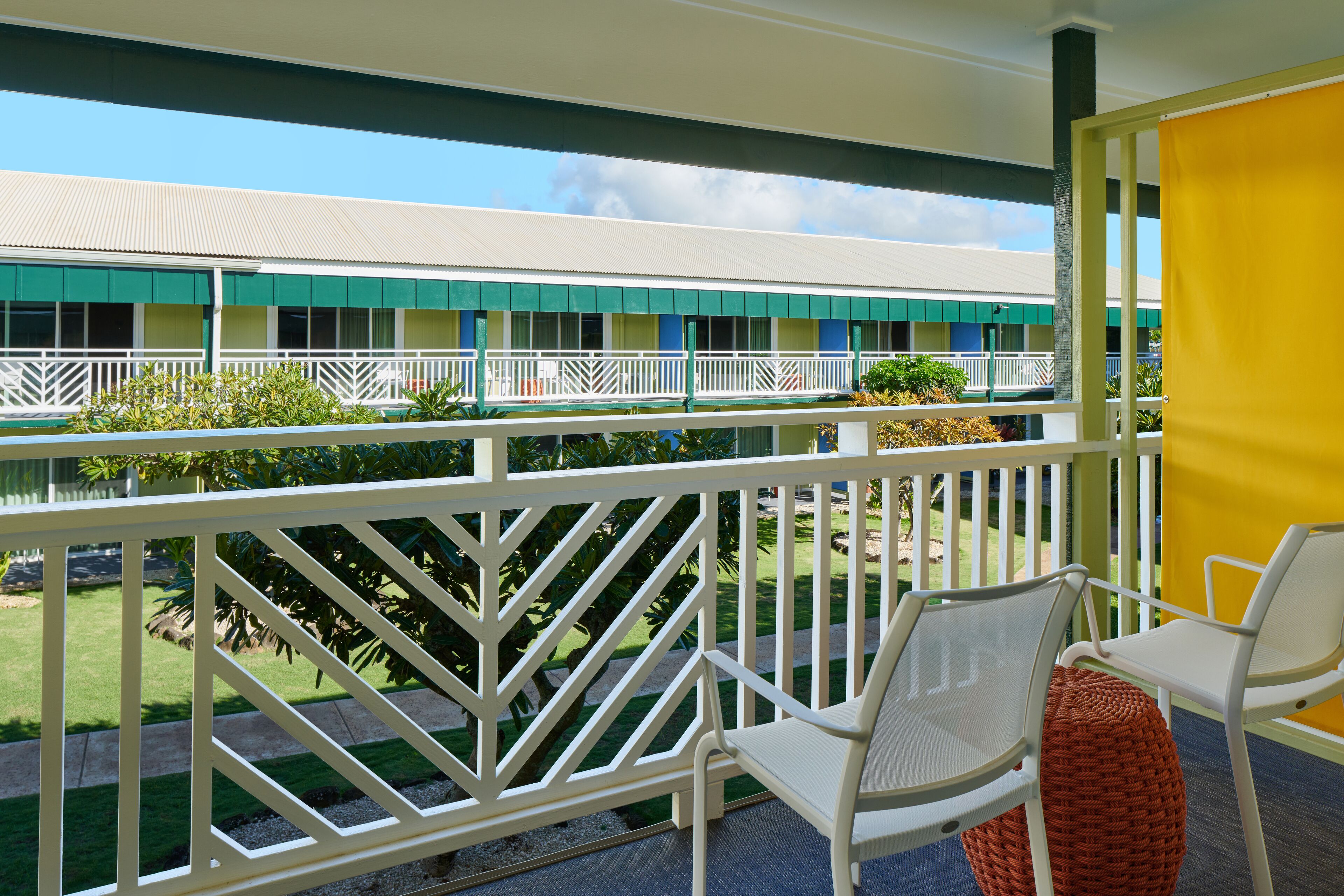 A balcony with white chairs and a yellow partition, overlooking a building with green and white exterior walls and a clear sky.