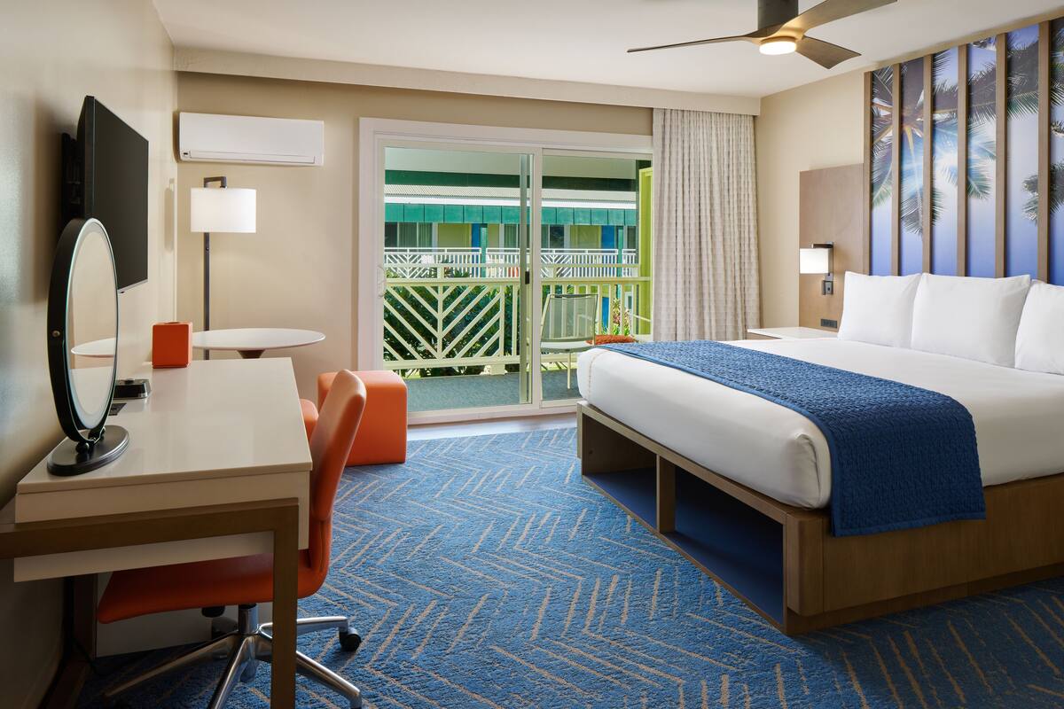 A hotel room with a bed, a desk with a chair, a small table, a mirror, and a balcony with a view of palm trees.