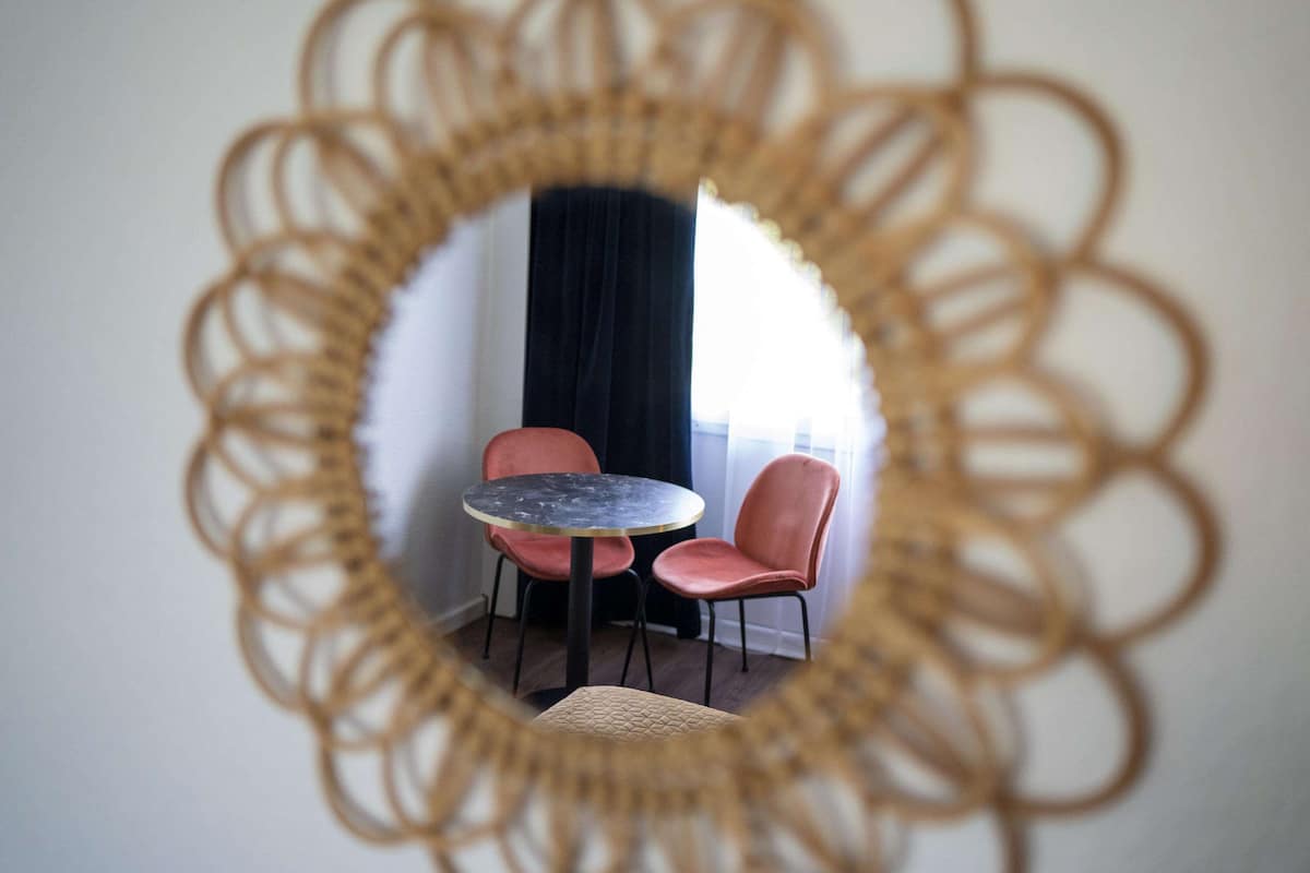 Um espelho redondo com moldura decorativa refletindo um quarto com uma mesa redonda e duas cadeiras.