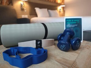 Habitació d'hotel amb llit, almoines, una estora de ioga, peses i un fullet informatiu de fitness.