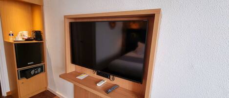 A modern hotel room with a large flat-screen TV mounted on a wooden wall, a small wooden shelf with a coffee machine, and a compact refrigerator.