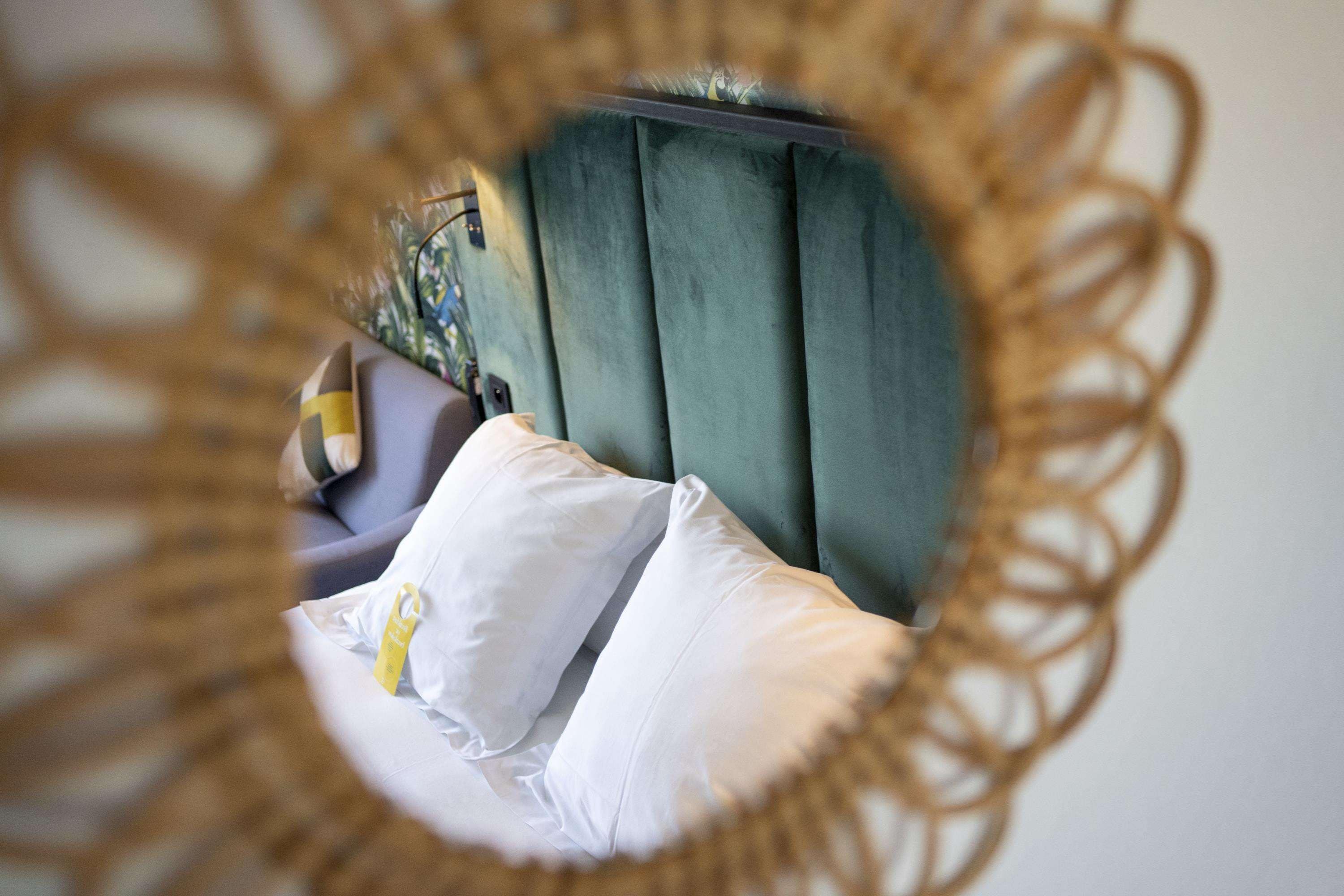 A round mirror with a decorative frame reflecting a room with a bed, pillows, and a green curtain.