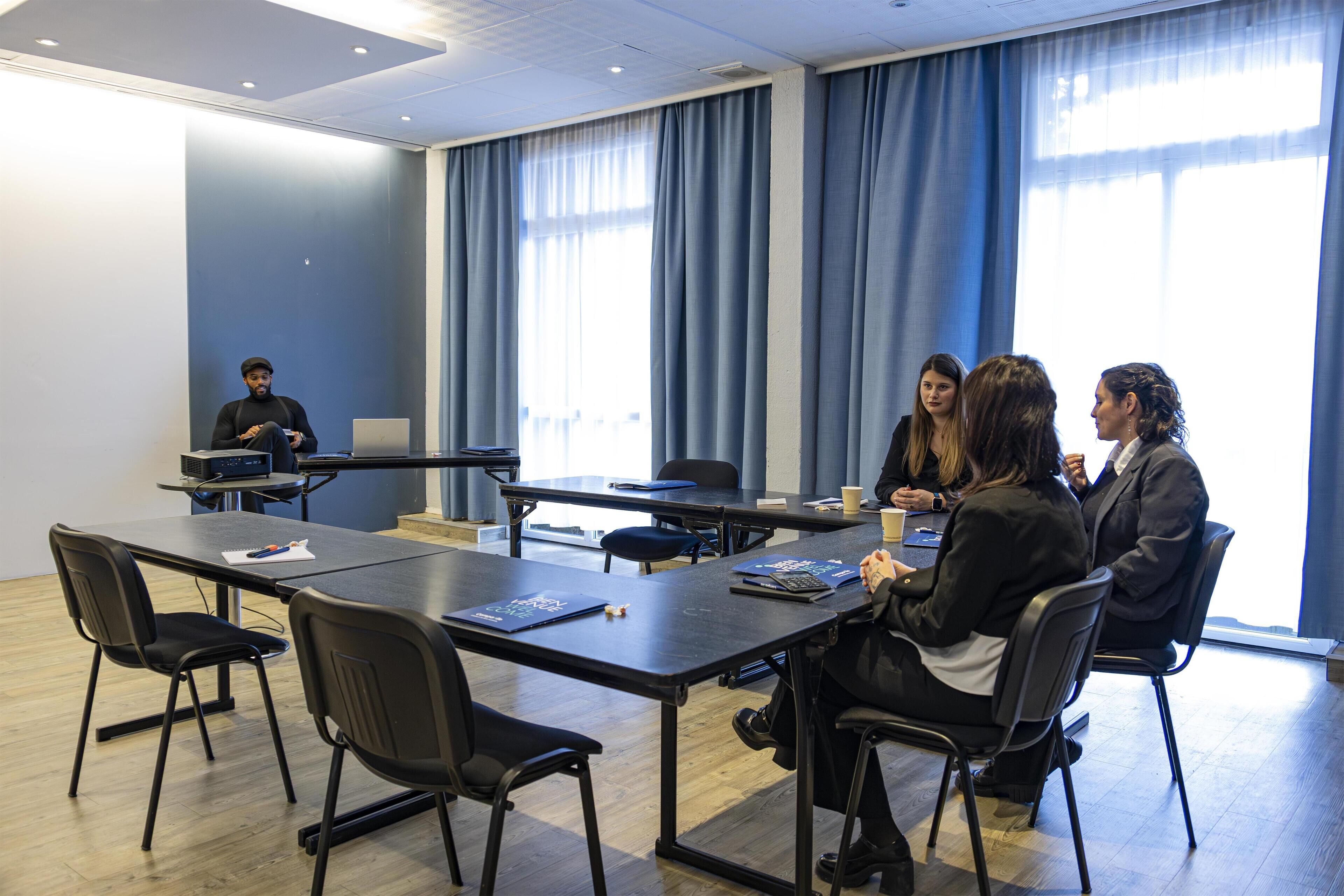 A conference room with a long table, black chairs, and a person standing at the front.