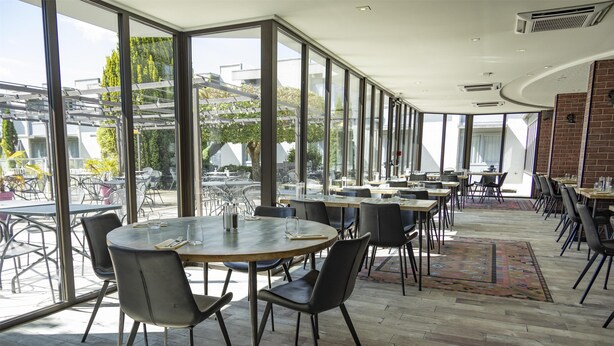 A modern dining area with round tables and chairs, large windows offering views of the outdoors, and a mix of natural and artificial lighting. - Campanile PRIME - Troyes Nord Barberey (Barberey-Saint-Sulpice)