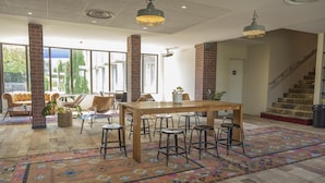 A spacious room with a large wooden table, multiple bar stools, and a colorful patterned rug. There are large windows allowing natural light, and a staircase is visible in the background. - Campanile PRIME - Troyes Nord Barberey (Barberey-Saint-Sulpice)