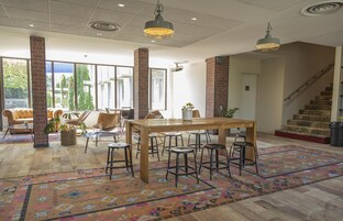 Una habitación amplia con una gran mesa de madera, varios taburetes y un tapete colorido con estampado. Hay ventanas grandes que permiten la entrada de luz natural, y se ve una escalera al fondo.
