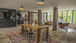 A spacious indoor area with a wooden table and bar stools, surrounded by large windows offering a view of greenery outside. - Campanile PRIME - Troyes Nord Barberey (Barberey-Saint-Sulpice)