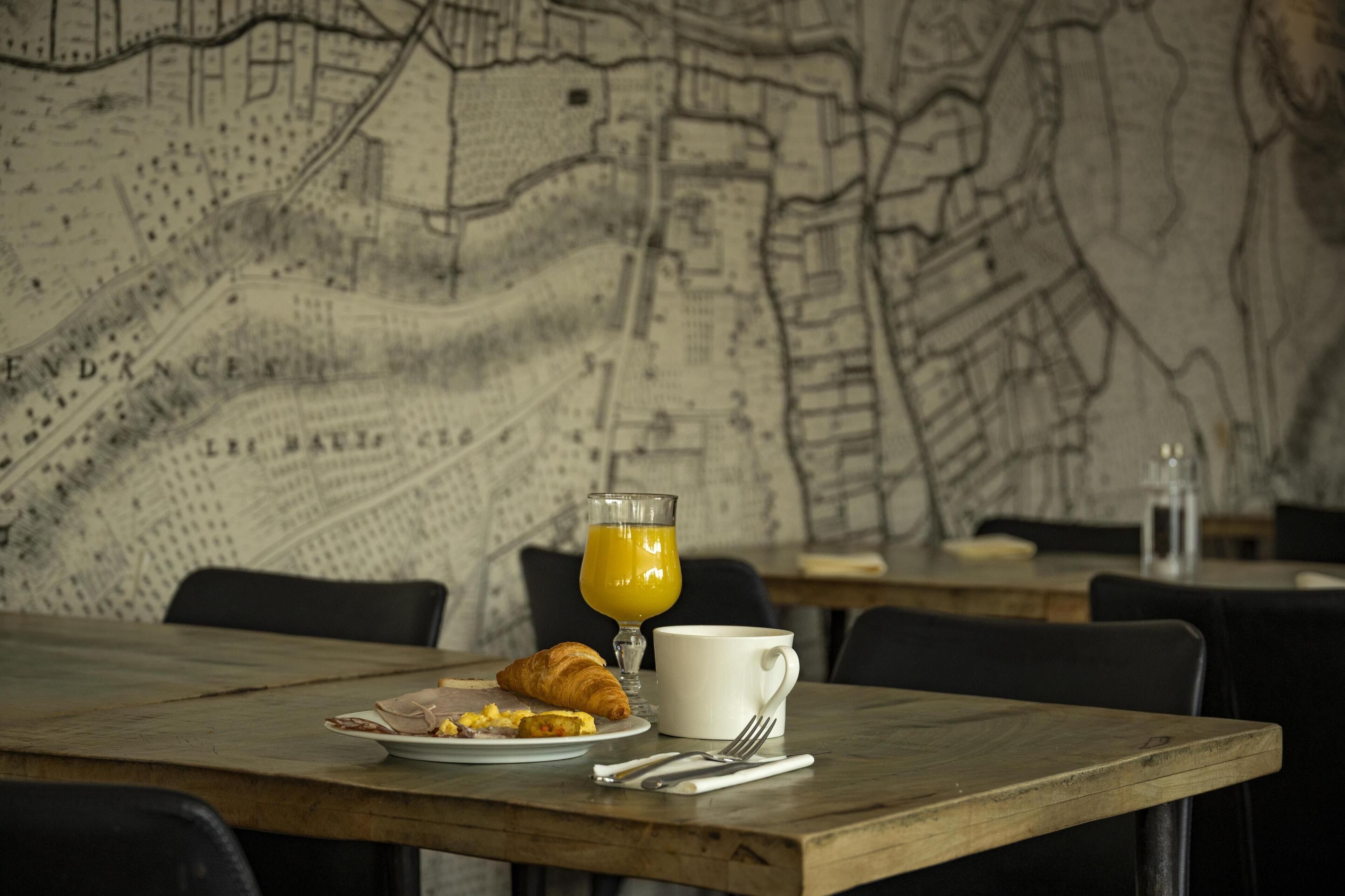 A breakfast setting with a map on the wall, a croissant, a glass of orange juice, and a white mug on a wooden table.