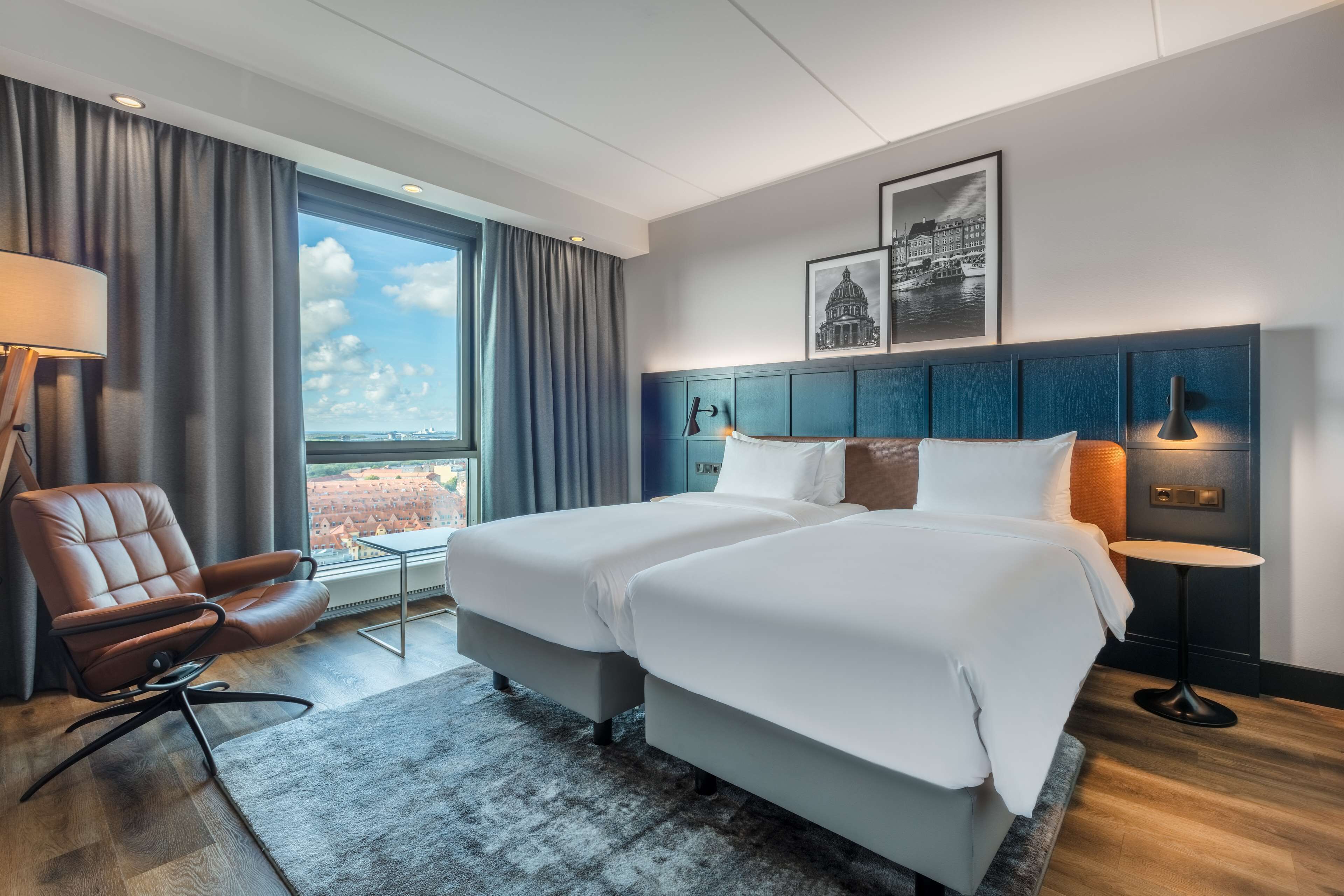 A hotel room with two beds, a desk, a chair, a window with curtains, and a cityscape view.