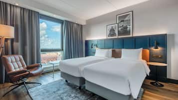 Habitación de hotel con dos camas, un escritorio, una silla, una ventana con cortinas y vistas a la ciudad.