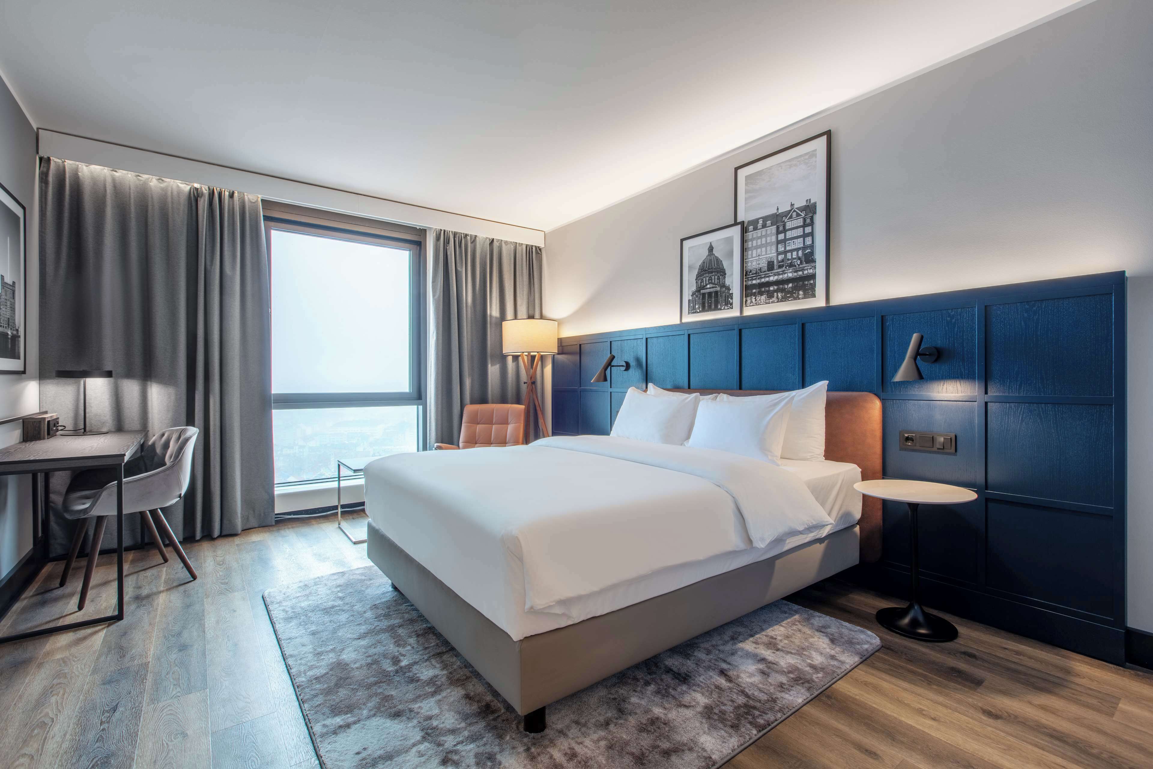 A modern hotel room with a large bed, a desk, and a view of the city.