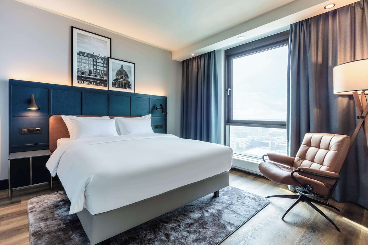 A hotel room with a large bed, a desk, a chair, and a view of the city through a large window.