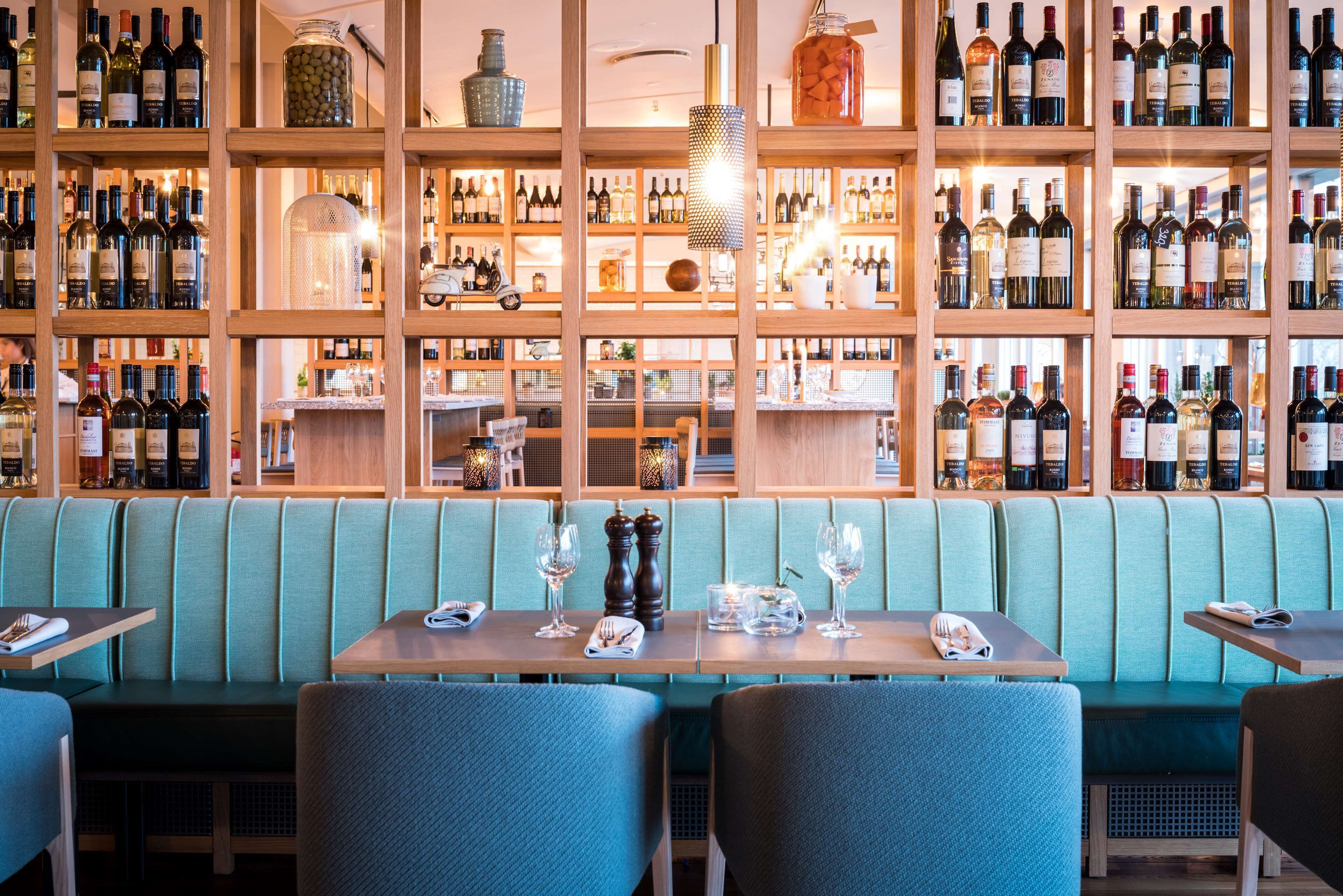 Un restaurante con estanterías de madera llenas de botellas de vino, una cabina acolchada de color verde y una mesa con copas de vino y cubiertos.