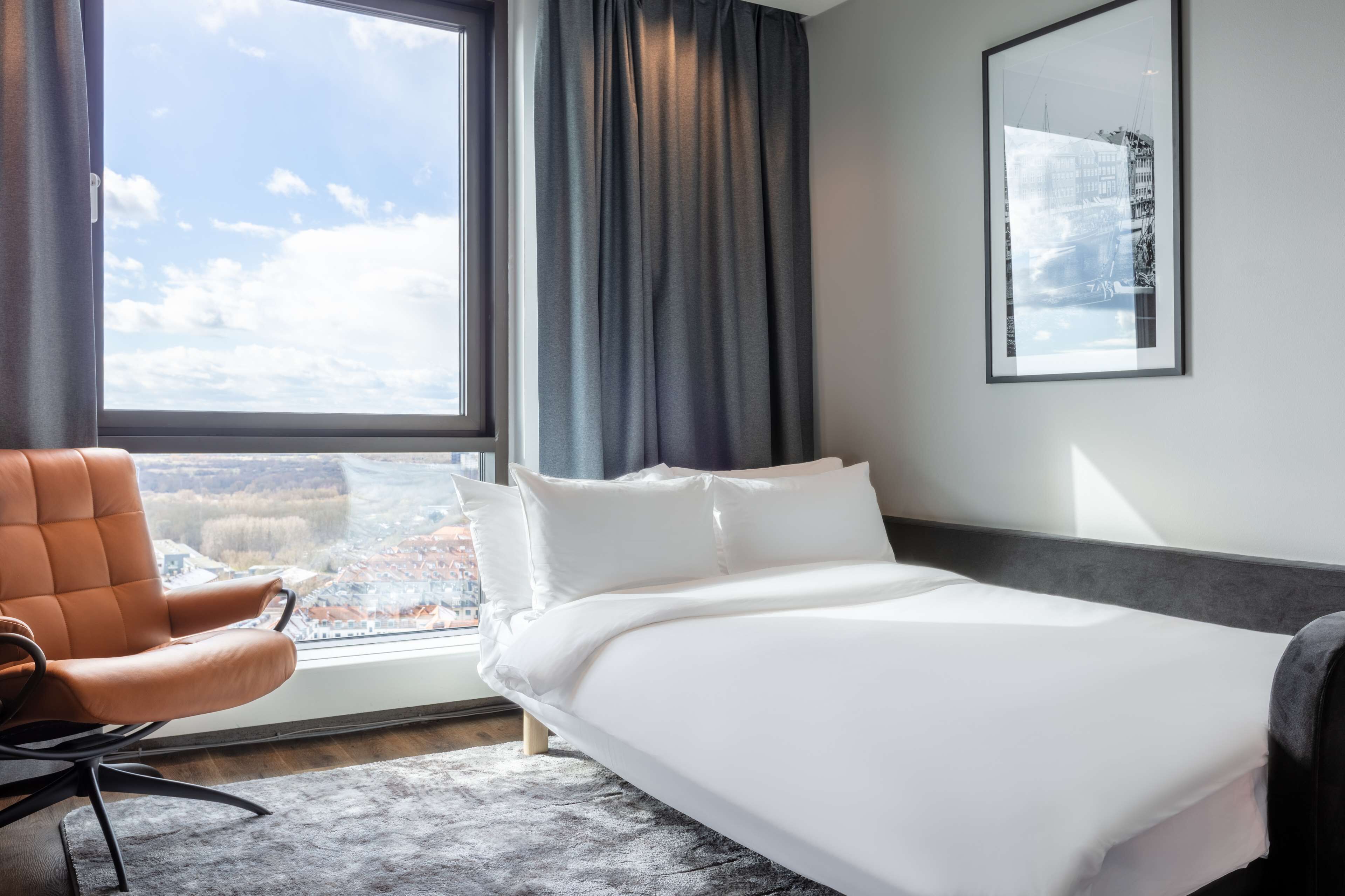 A hotel room with a bed, a chair, a window with a view of a cityscape, and a framed picture on the wall.