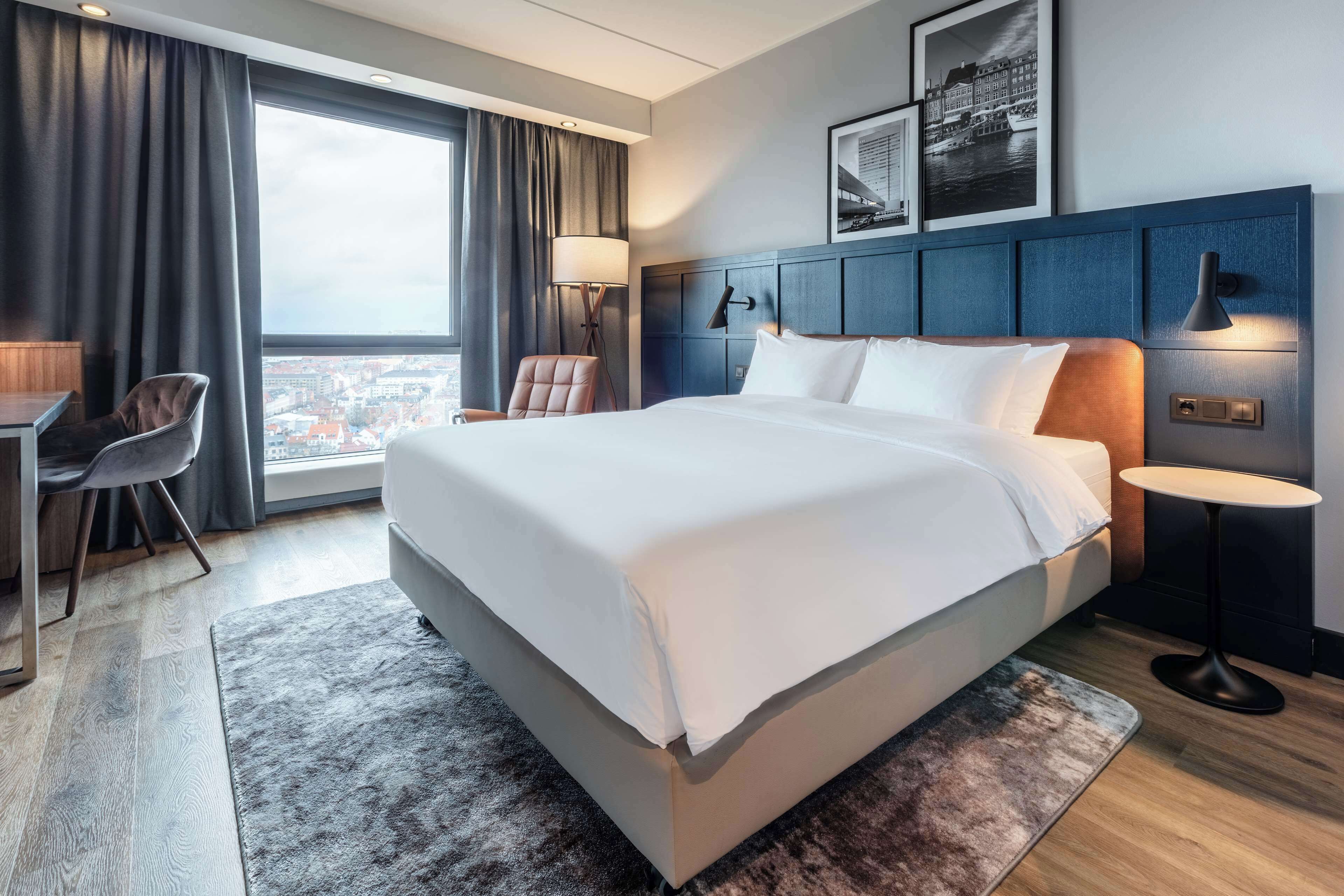 A modern hotel room with a large bed, a desk, a chair, and a view of the city through a large window.