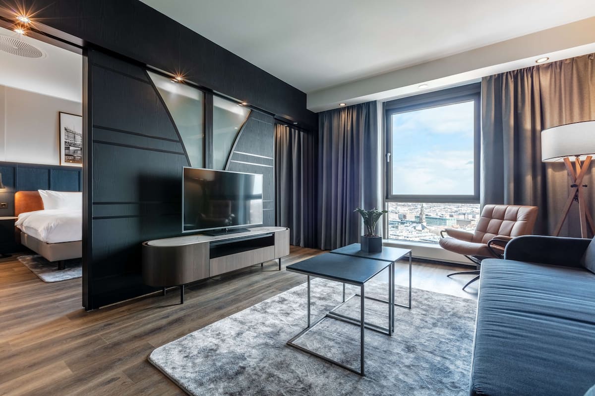 A modern hotel room with a large window, a flat-screen TV, a sofa, a coffee table, and a bed with a headboard.