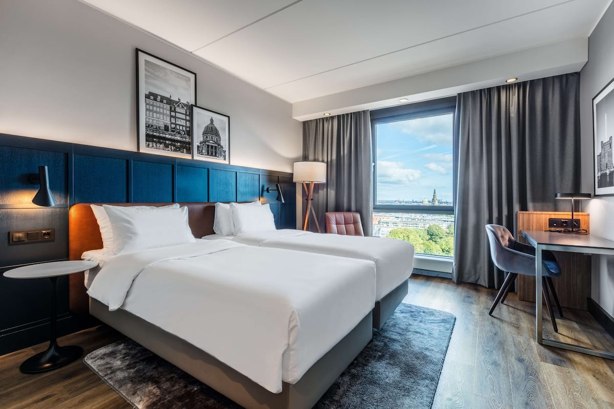 A hotel room with two beds, a desk, a chair, a lamp, and a view of the city.