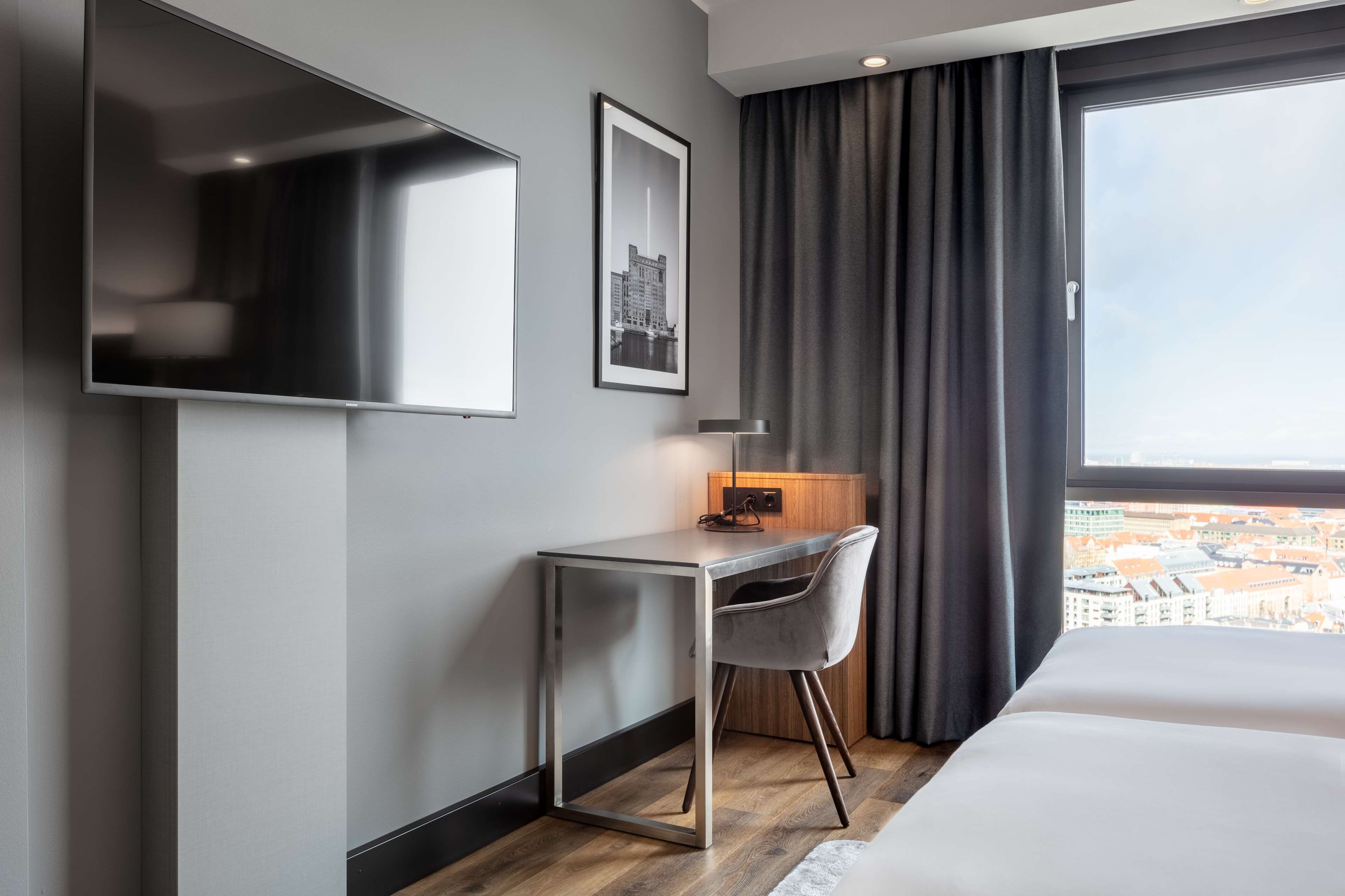Une chambre d’hôtel moderne équipée d’un lit, d’un bureau, d’une chaise, d’un téléviseur à écran plat, d’un tableau encadré et d’une fenêtre donnant sur la ville.