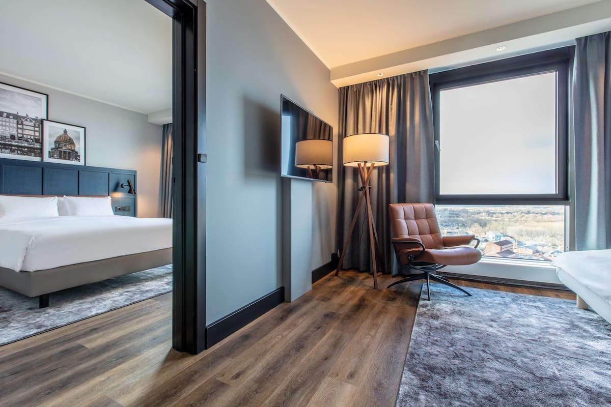 A hotel room with a bed, a chair, a lamp, a large window, and a view of the city.