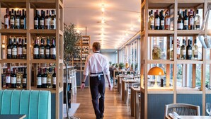 Un restaurant avec des étagères en bois remplies de bouteilles de vin, un serveur qui se déplace et un banc recouvert d’un tissu turquoise.
