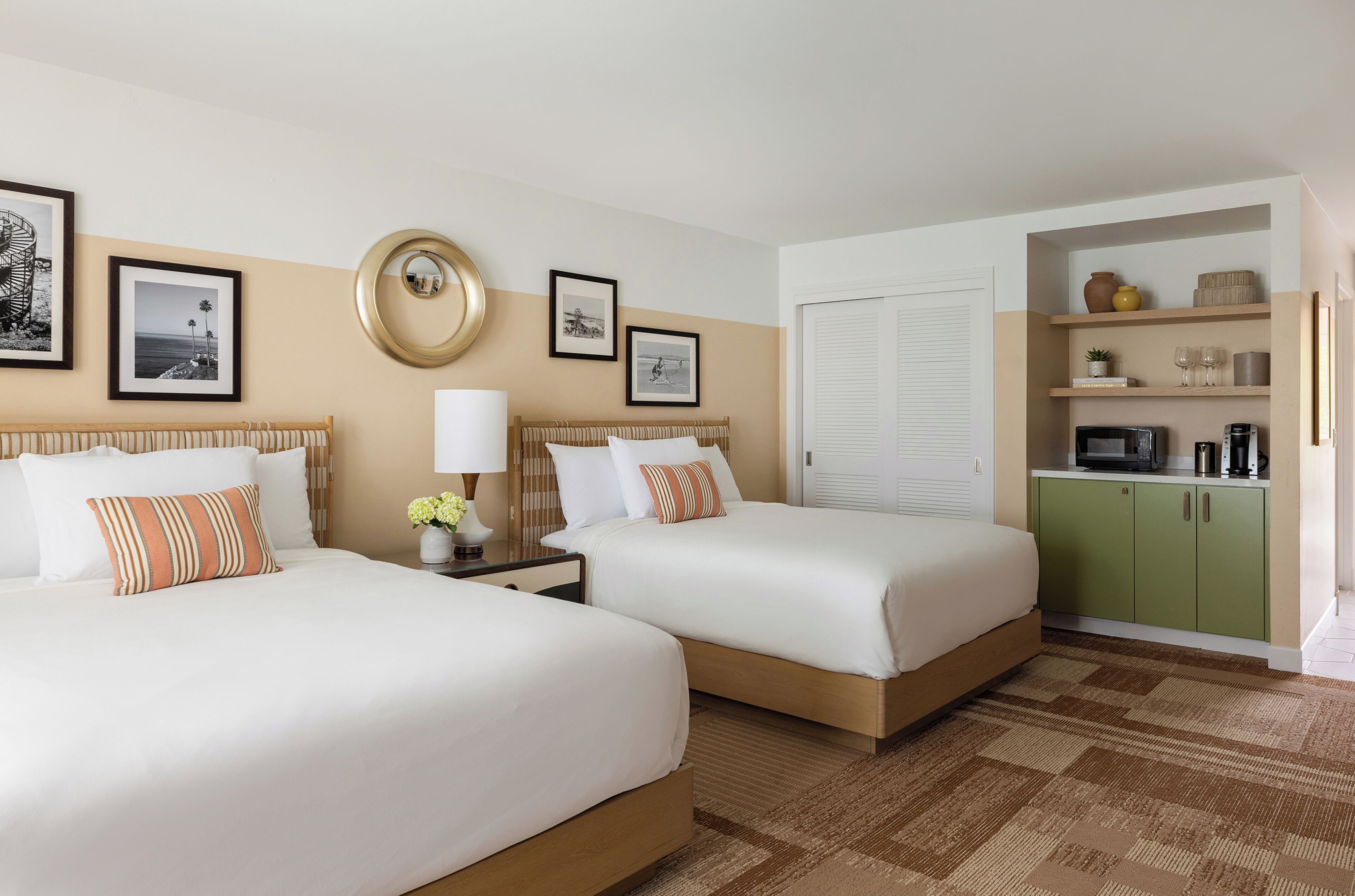 A hotel room with two beds, a bedside table with a lamp, a wall-mounted mirror, framed pictures, a green minibar, and a window with closed blinds.