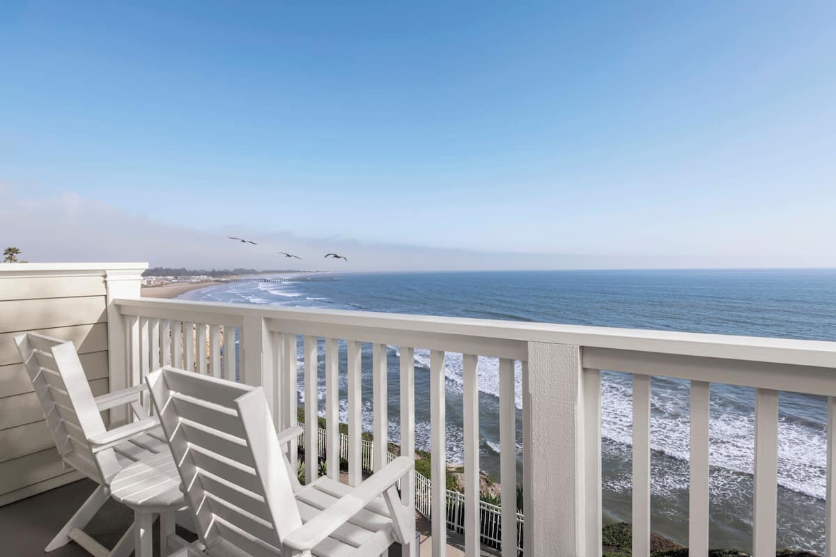 A balcony with white railings overlooking the ocean, featuring two white Adirondack chairs.