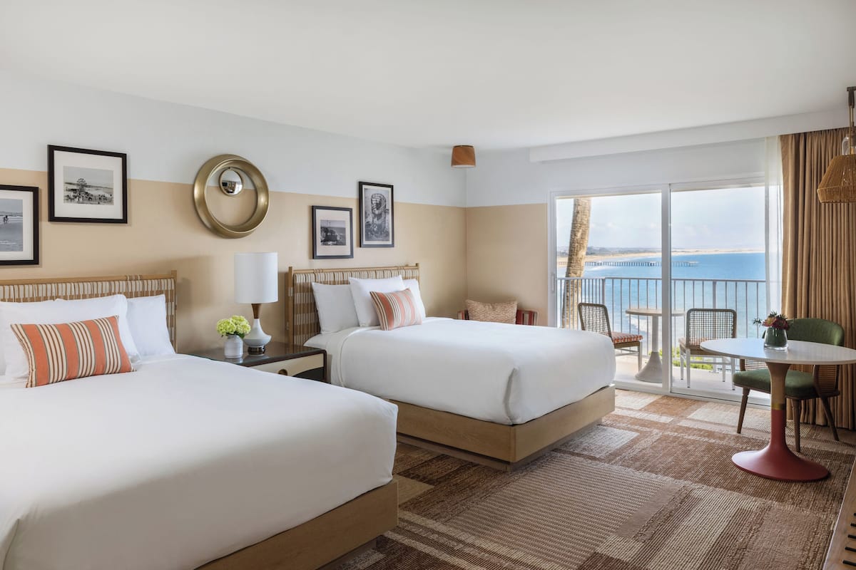 A hotel room with two beds, a balcony with a view of the sea, and a dining area with a table and chairs.