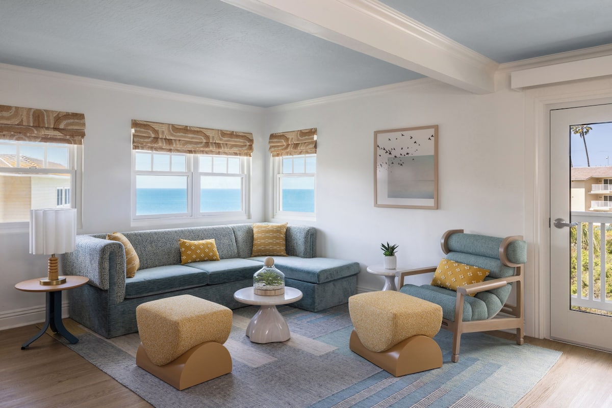 A living room with a sectional sofa, a round coffee table, and a chair. There are windows with patterned valances, a framed picture on the wall, and a door with a view of palm trees.