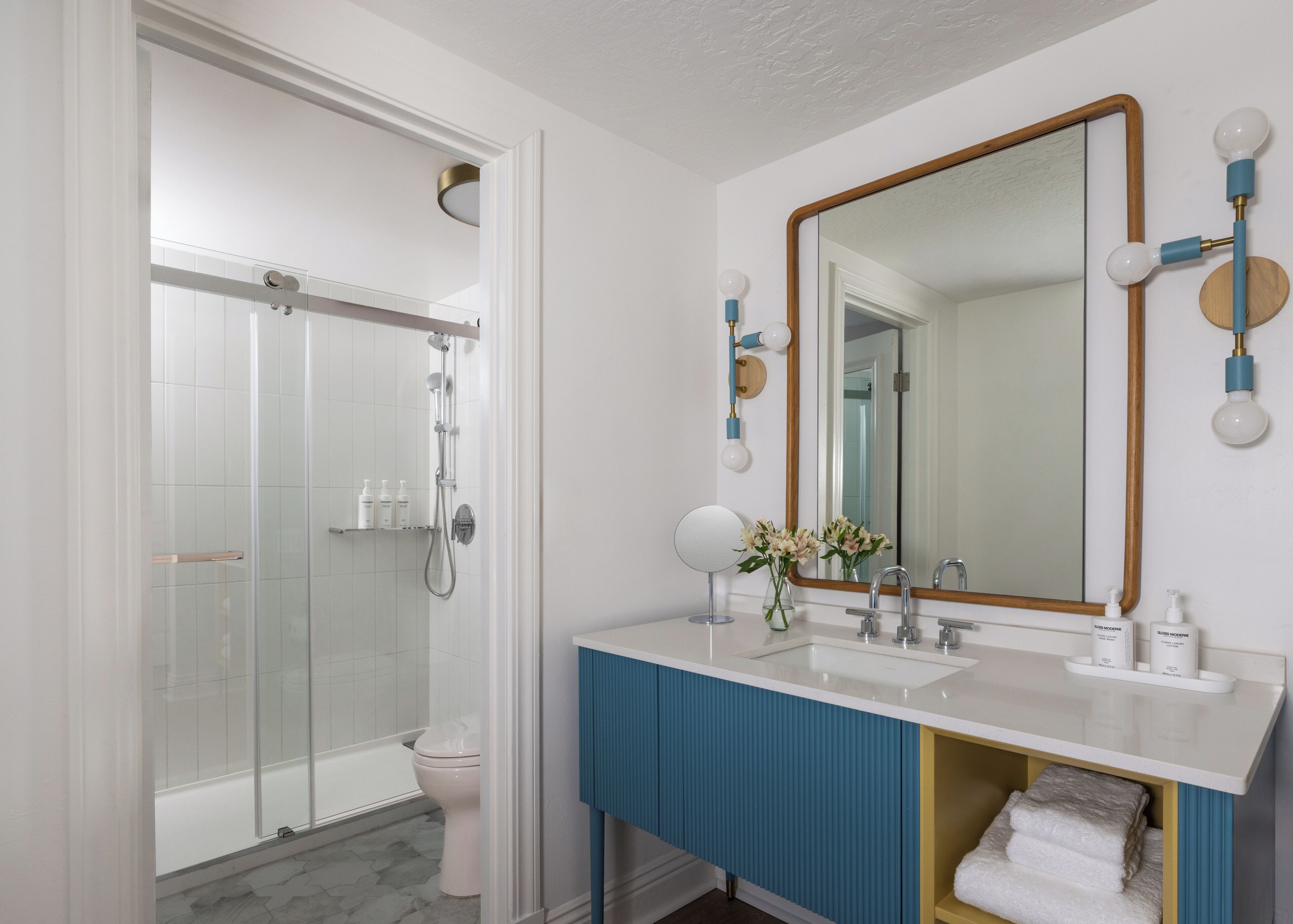 A modern bathroom with a glass-enclosed shower, a white sink with blue accents, and a large mirror with round lights.