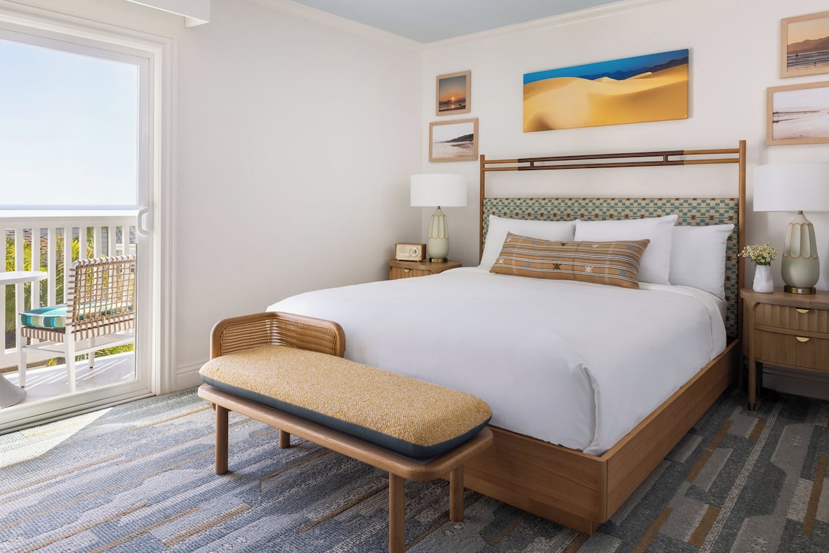 A hotel room with a large bed, a bench, a nightstand, a lamp, and a view of the outdoors.