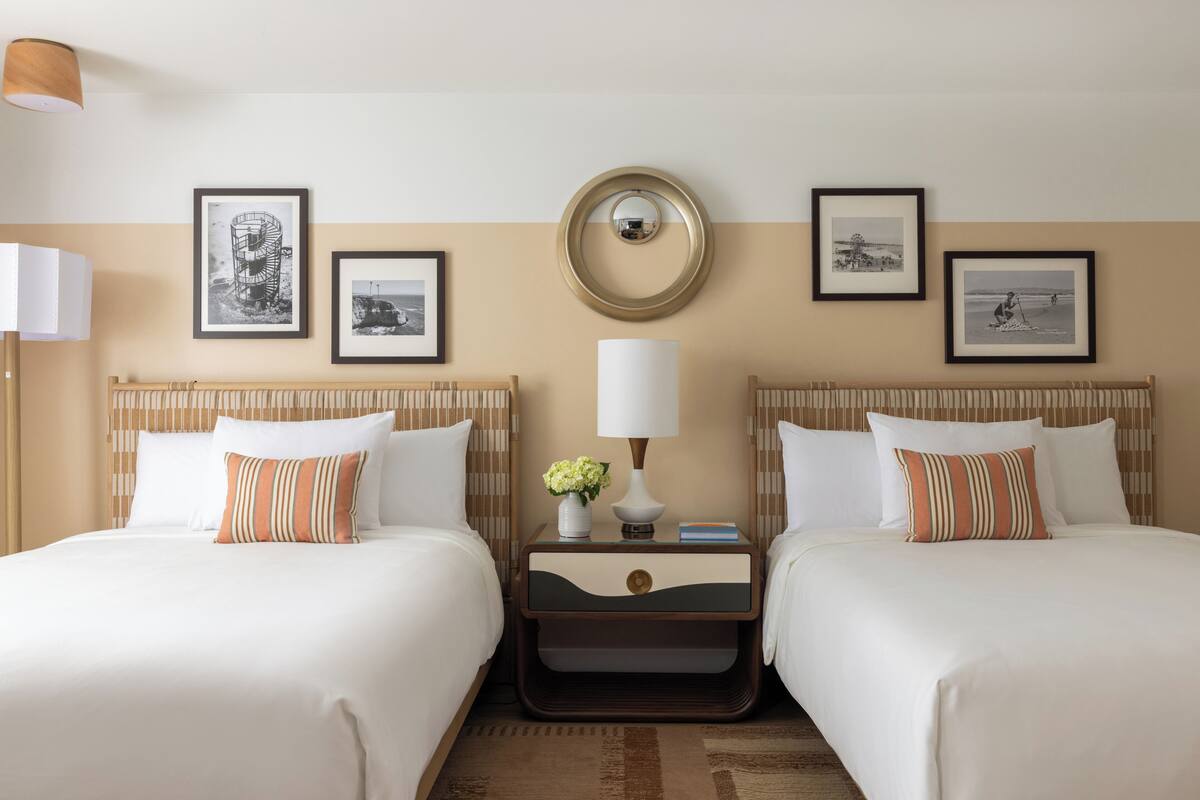 A hotel room with two beds, a nightstand with a lamp, framed pictures on the wall, and a wooden headboard.