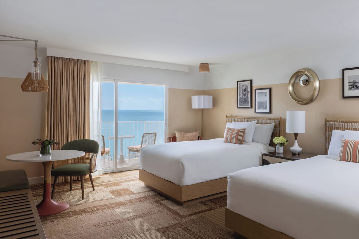 A hotel room with two beds, a dining area, and a balcony with a view of the sea.