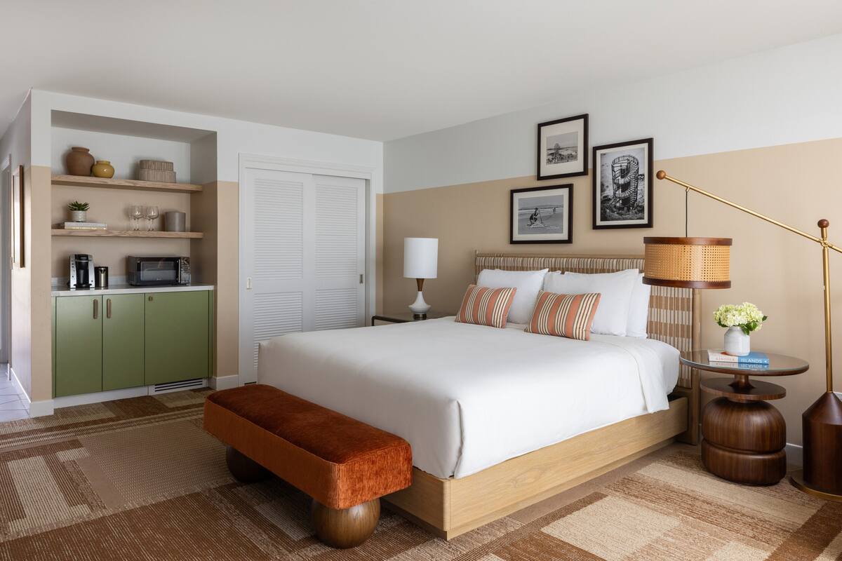 A bedroom with a large bed, a bench, a bedside table, a lamp, and a shelf with items.