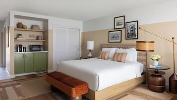 A hotel room with a large bed, a bench, a bedside table with a lamp, a wall-mounted shelf with items, and a door.