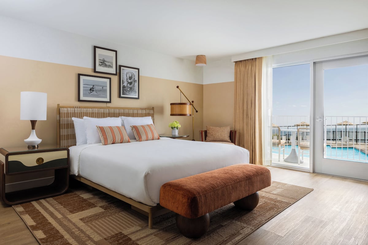 A hotel room with a large bed, a bedside table with a lamp, a small table with a vase, a bench, and a view of a pool through a sliding door.