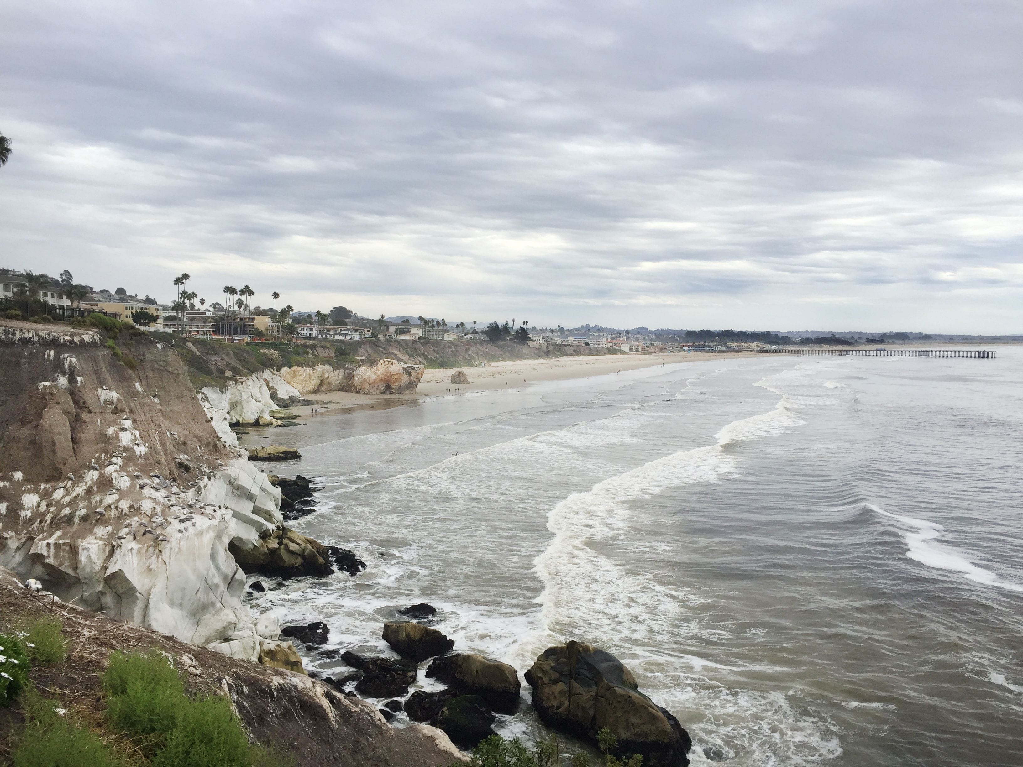 A coastal beach with rocky cliffs, a sandy shore, and a pier extending into the water.