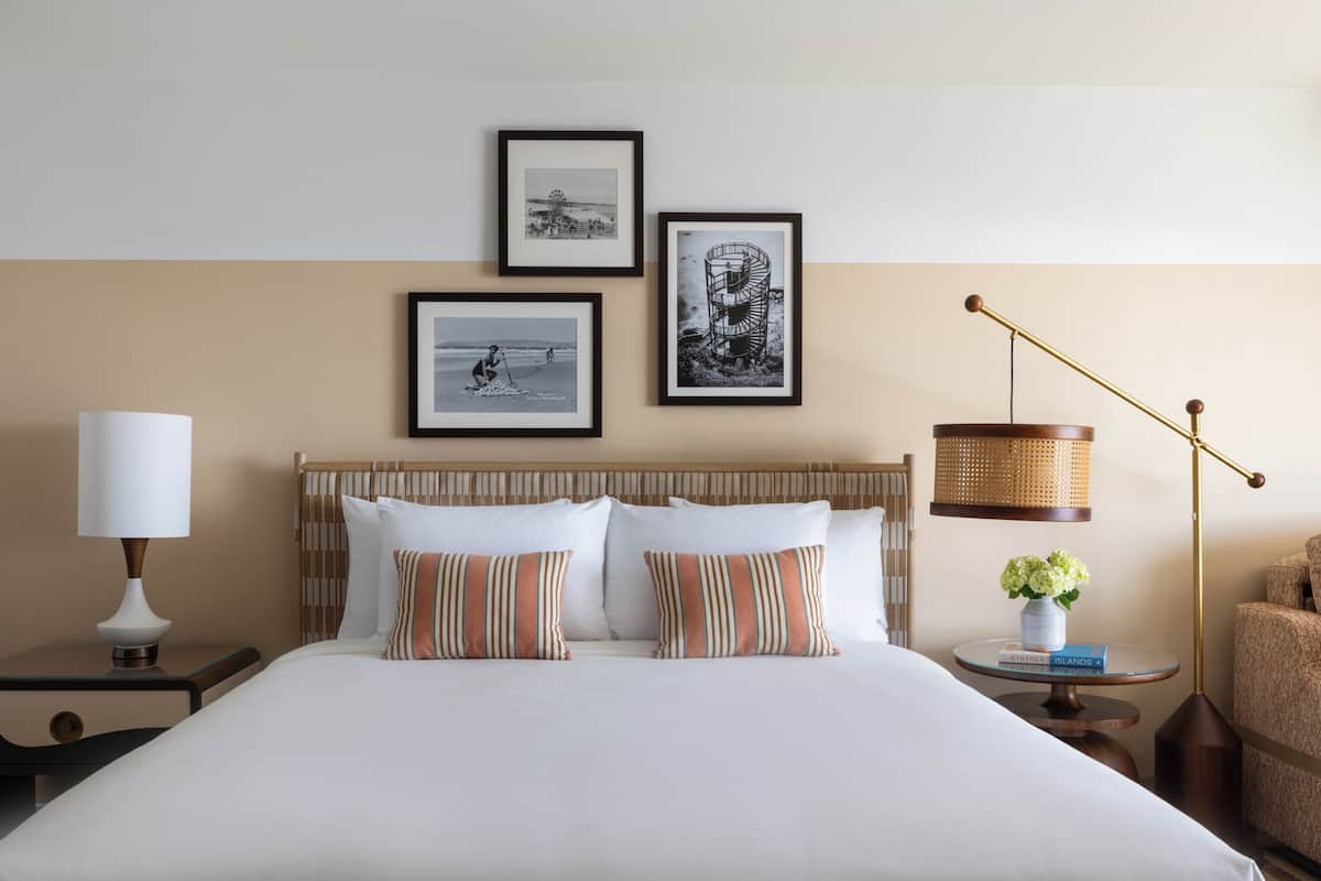 A hotel room with a bed, a nightstand, a lamp, framed pictures on the wall, and a small table with a vase and books.